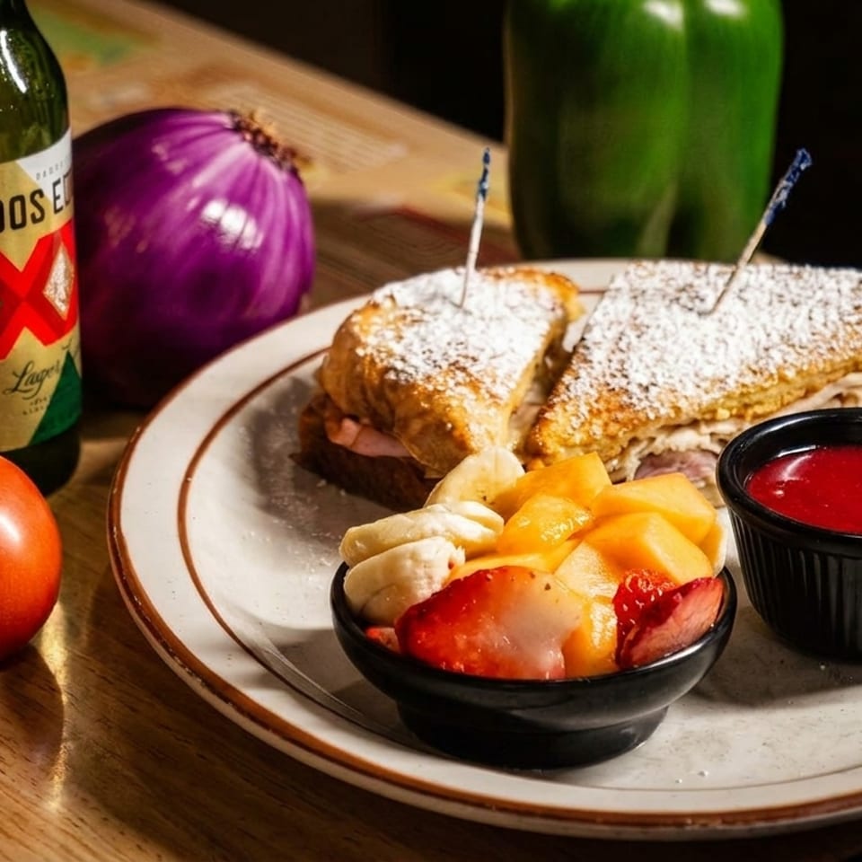 Grilled sandwich with powdered sugar, fruit bowl, and red dipping sauce beside Dos Equis beer, tomato, and onion on a wooden table.